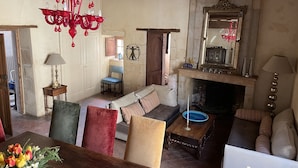 Ein gemütliches Wohnzimmer mit Kamin, einer Chandelierleuchtre und einem Spiegel. Es gibt ein Sofa, einen Tisch und einen Sessel.