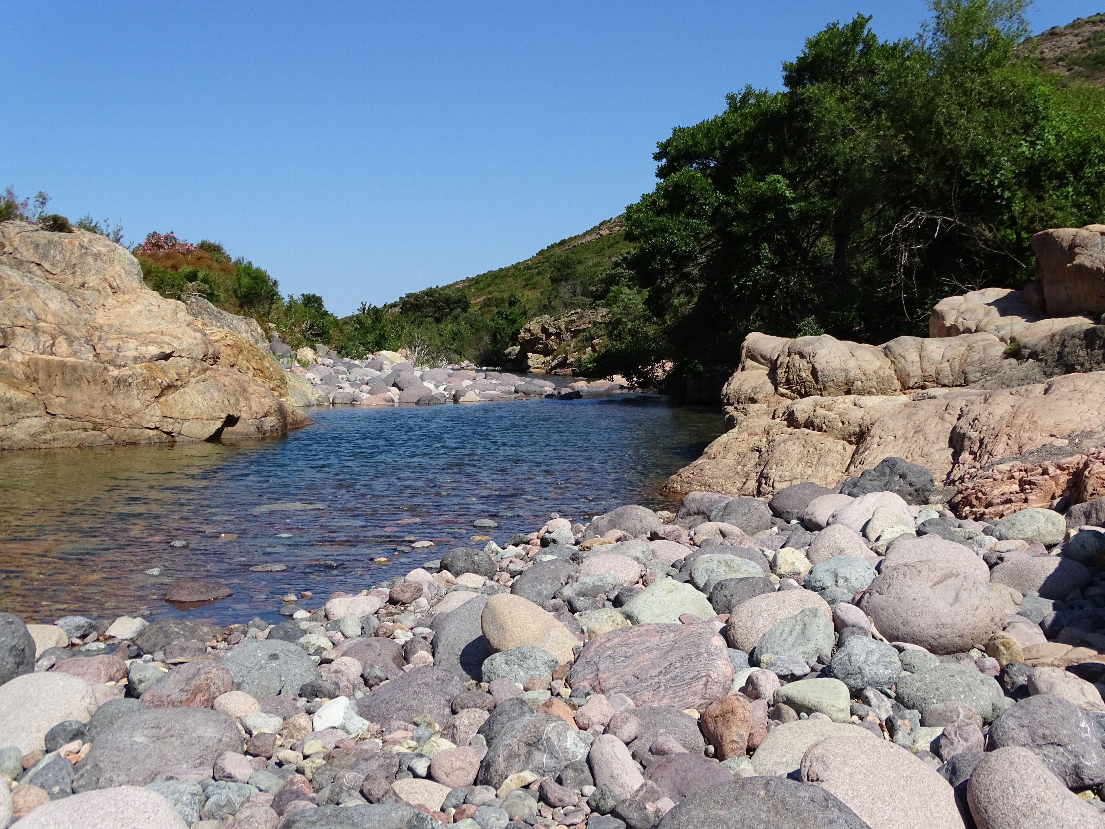 Orilla rocosa de río con agua cristalina, rodeada de vegetación y grandes rocas.