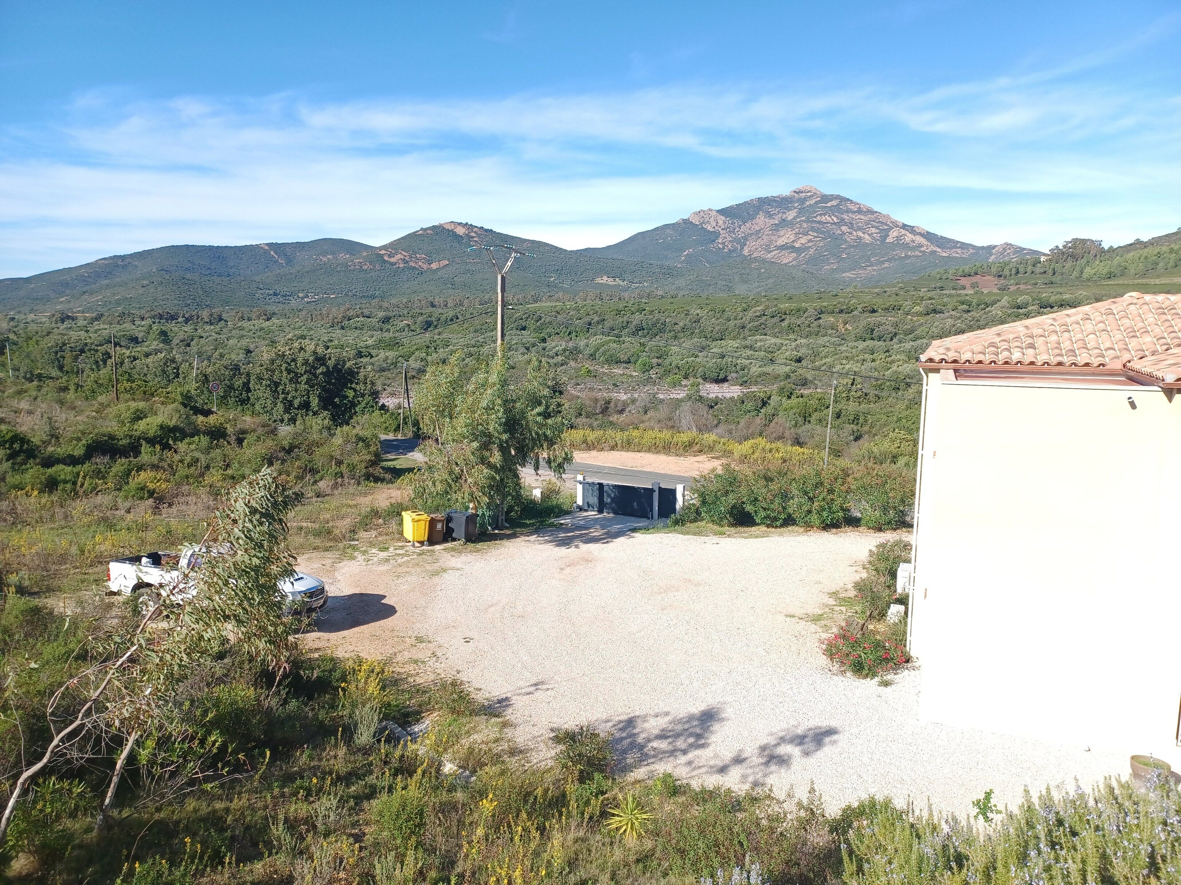 A rural setting with a gravel driveway, a small building with a tiled roof, and a mountain in the background.