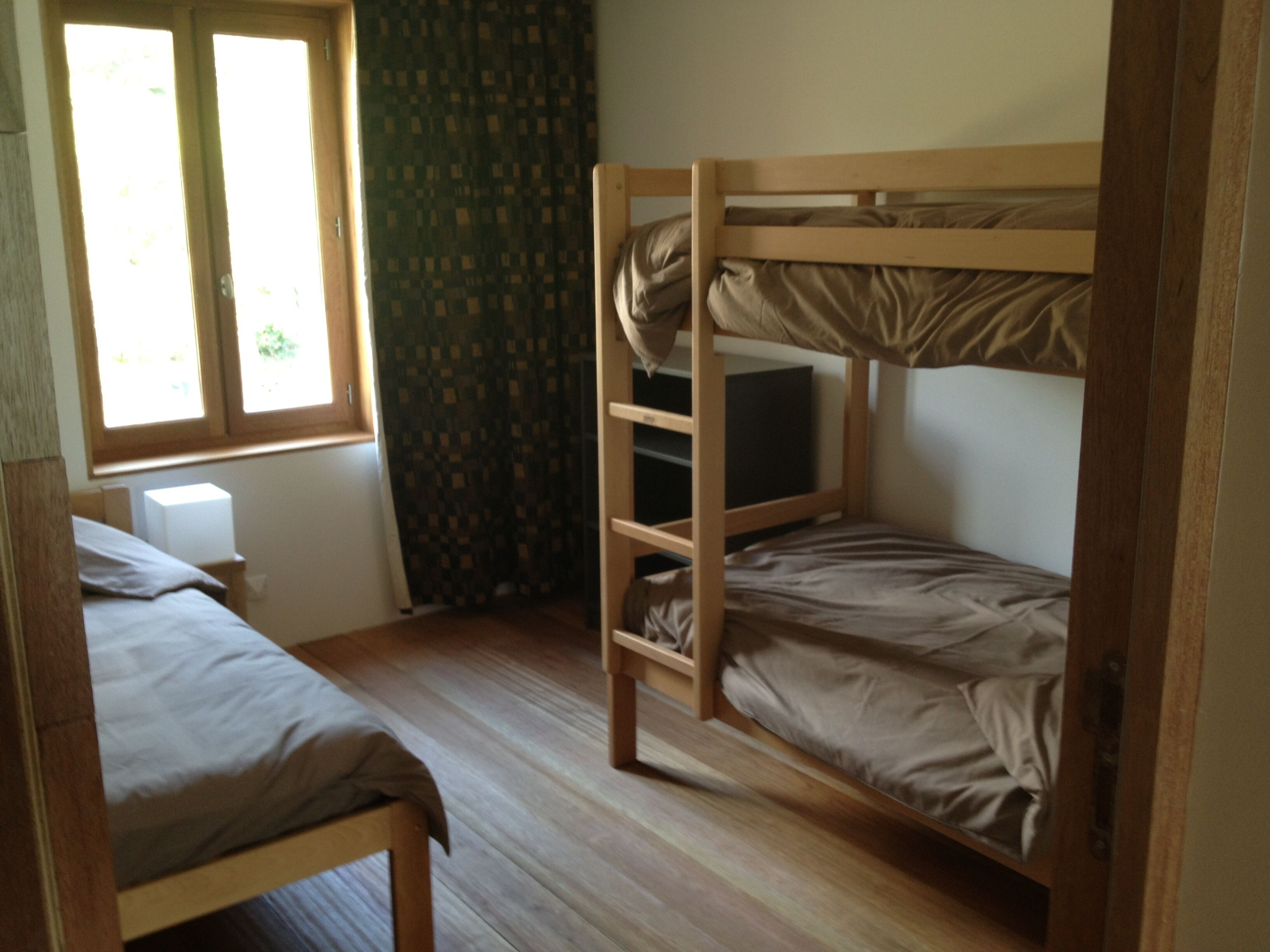 A room with two bunk beds, a window with curtains, and a small bedside table with a lamp.