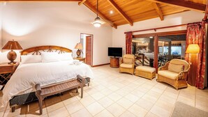 Ruime slaapkamer met een groot bed, twee rieten fauteuils, een televisie en een schuifdeur naar een balkon.