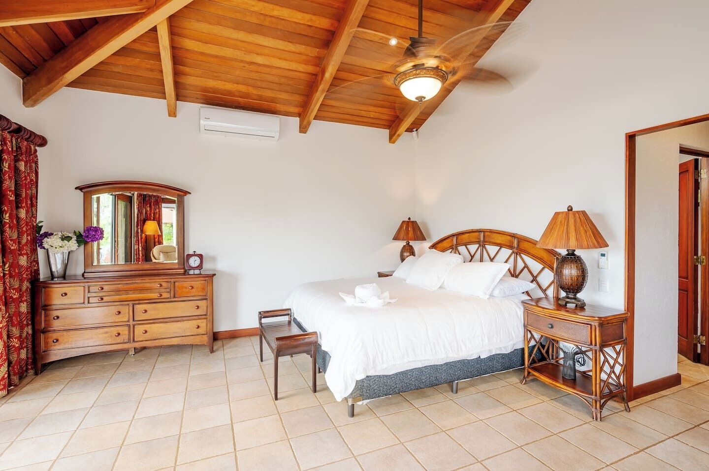 A bedroom with a wooden ceiling, a bed with white bedding, a wooden dresser, a mirror, two bedside lamps, and a small table with a lamp.