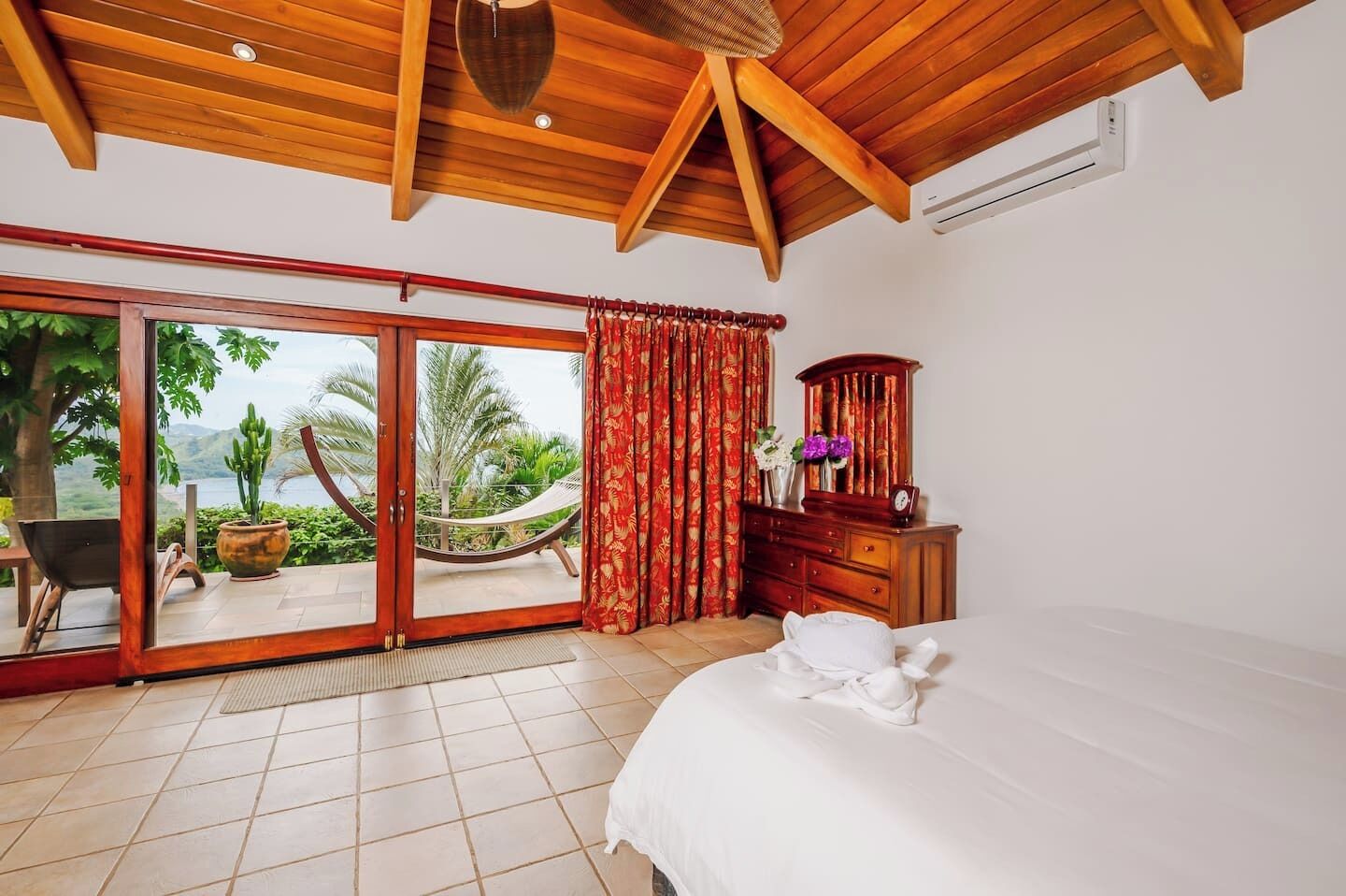 A bedroom with a large bed, a wooden dresser, a nightstand, a red curtain, and a view of the outdoors.