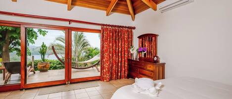 A bedroom with a large bed, a wooden dresser, a nightstand, a red curtain, and a view of the outdoors.