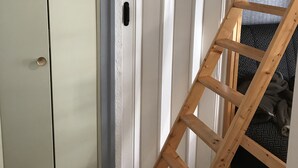 A wooden ladder leaning against a white door with a small hole, possibly for ventilation, and a patterned fabric visible in the background. - L'Idéal en Charteuse (Saint-Pierre-de-Chartreuse)