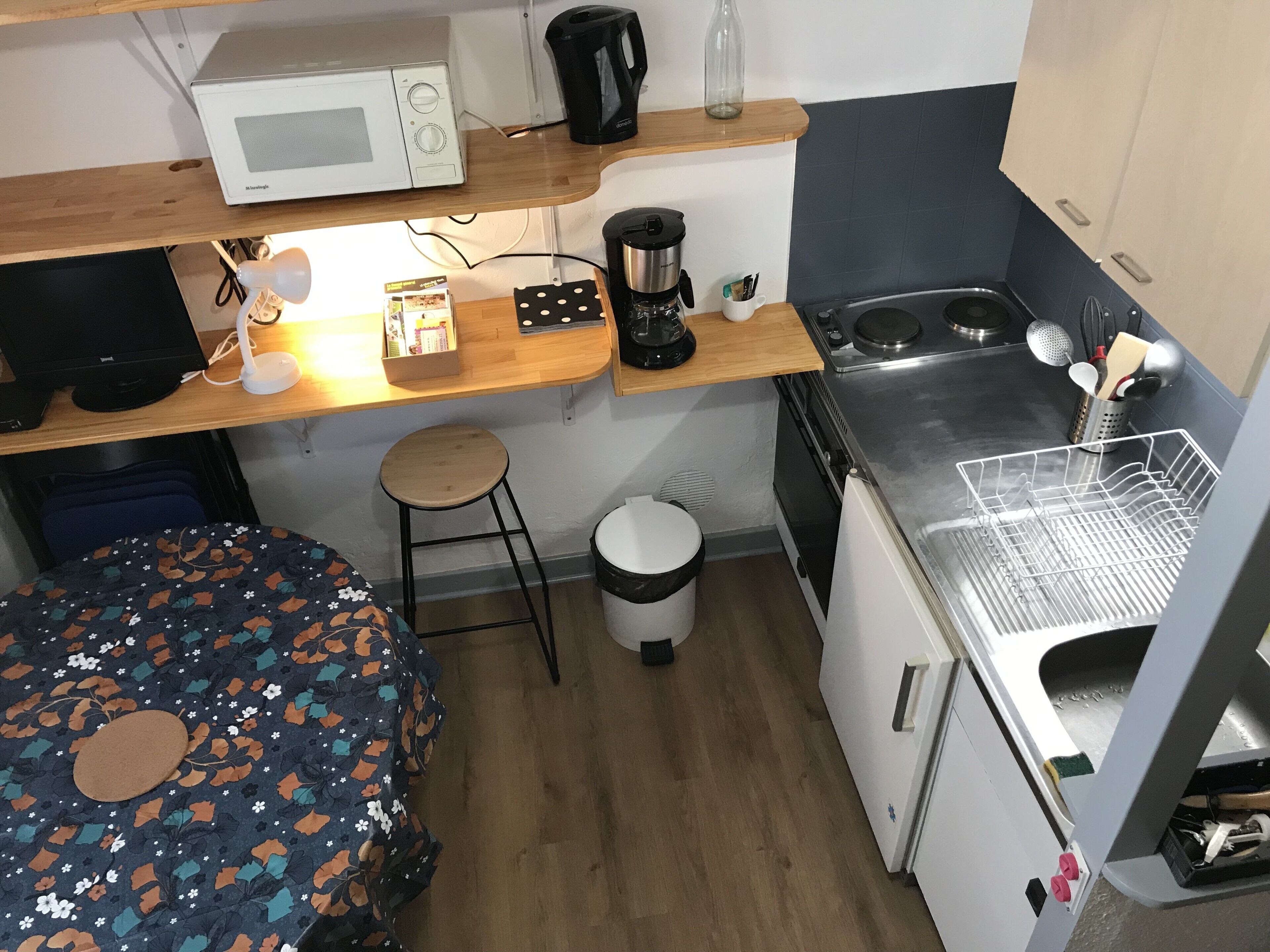 A compact kitchenette with a microwave, coffee maker, and stove. There's a small stool and a trash bin.