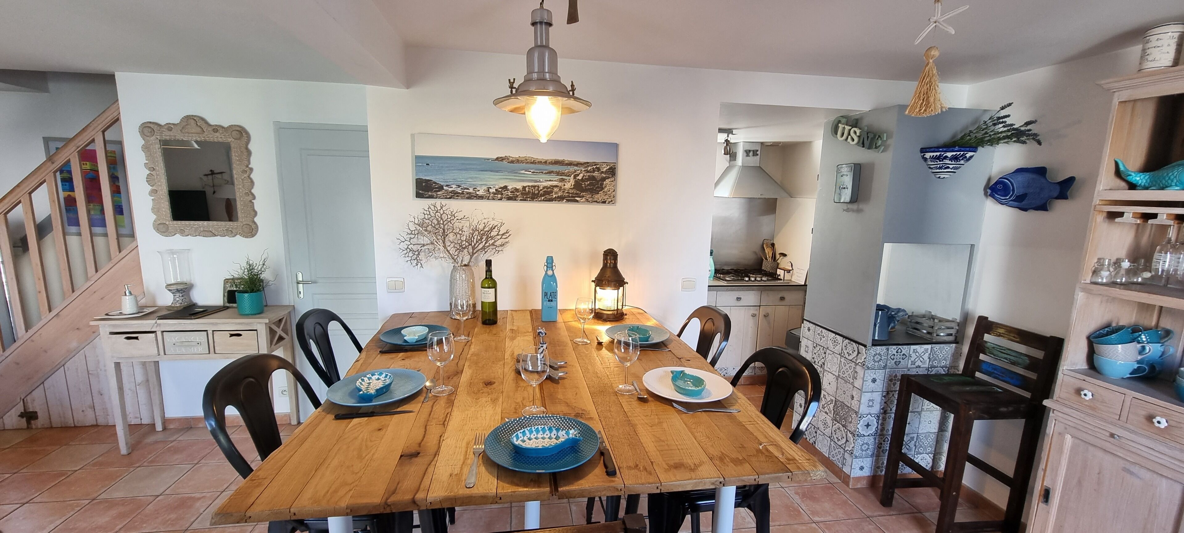Une salle à manger avec une table en bois dressée pour six personnes, entourée de chaises. Un escalier est visible sur la gauche, et une cuisine se trouve au fond de la pièce.