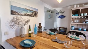 Un coin repas avec une table en bois dressée pour quatre personnes, une épée décorative accrochée au mur et un tableau représentant un paysage côtier au-dessus de la table.
