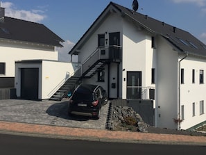 A modern two-story house with a car parked in the driveway, a staircase leading to the entrance, and a satellite dish on the roof. - Apartment Pellenzblick ground floor 92 m² (Nickenich)