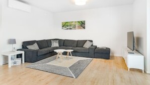 A modern living room with a dark grey sectional sofa, a small white side table with a lamp, a coffee table, and a flat-screen TV on a white stand.