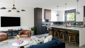 A modern kitchen with a central island, bar stools, a flat-screen TV, and a seating area with a sofa and chairs. - Gated Luxe Home with Rooftop, Bunk Room, & Parking  |  Sleeps 10 with King Suite, Queen Rooms & Custom Bunks, Chef’s Kitchen w/ Wine Fridge,  Rooftop Patio, Dog Friendly, Dedicated Parking, Mins from Downtown and Broadway, 24 Hour Hospitality (Nashville)