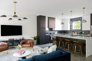 A modern kitchen with a central island, bar stools, a flat-screen TV, and a seating area with a sofa and chairs.