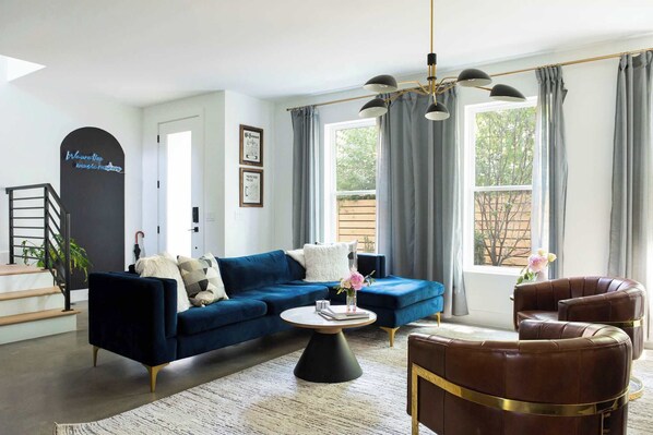 Una sala de estar moderna con un sofá azul, una mesa de centro redonda y un sillón de cuero. Se aprecia una escalera con barandilla, un ventanal con cortinas y una pared negra con un letrero de neón.