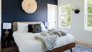 A bedroom with a large bed, a round wall decoration, two bedside lamps, and a window with a view of trees. - Gated Luxe Home with Rooftop, Bunk Room, & Parking  |  Sleeps 10 with King Suite, Queen Rooms & Custom Bunks, Chef’s Kitchen w/ Wine Fridge,  Rooftop Patio, Dog Friendly, Dedicated Parking, Mins from Downtown and Broadway, 24 Hour Hospitality (Nashville)