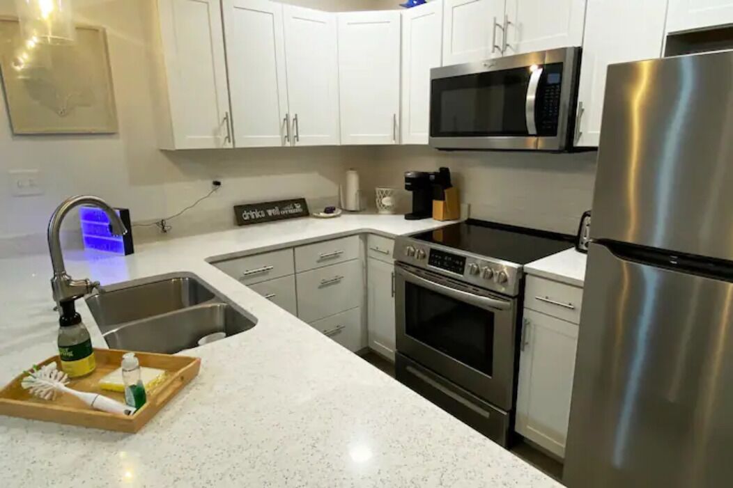 A modern kitchen with white cabinets, stainless steel appliances including a microwave, oven, and refrigerator, a sink with a faucet, and a tray with cleaning supplies.