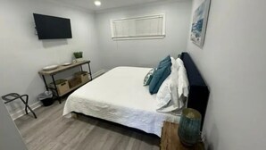 Una camera da letto con un letto, un comodino, una TV a parete, una finestra con tende e un piccolo ripiano con oggetti.