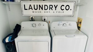 Un'area lavanderia con due lavatrici bianche, una ventola e un cartello che recita “LAUNDRY CO. WASH, DRY, & FOLD”.