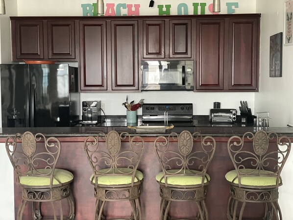 Una cocina con armarios de madera oscura, un refrigerador negro, una cafetera, un tostador y un taburete decorativo con cojín verde.