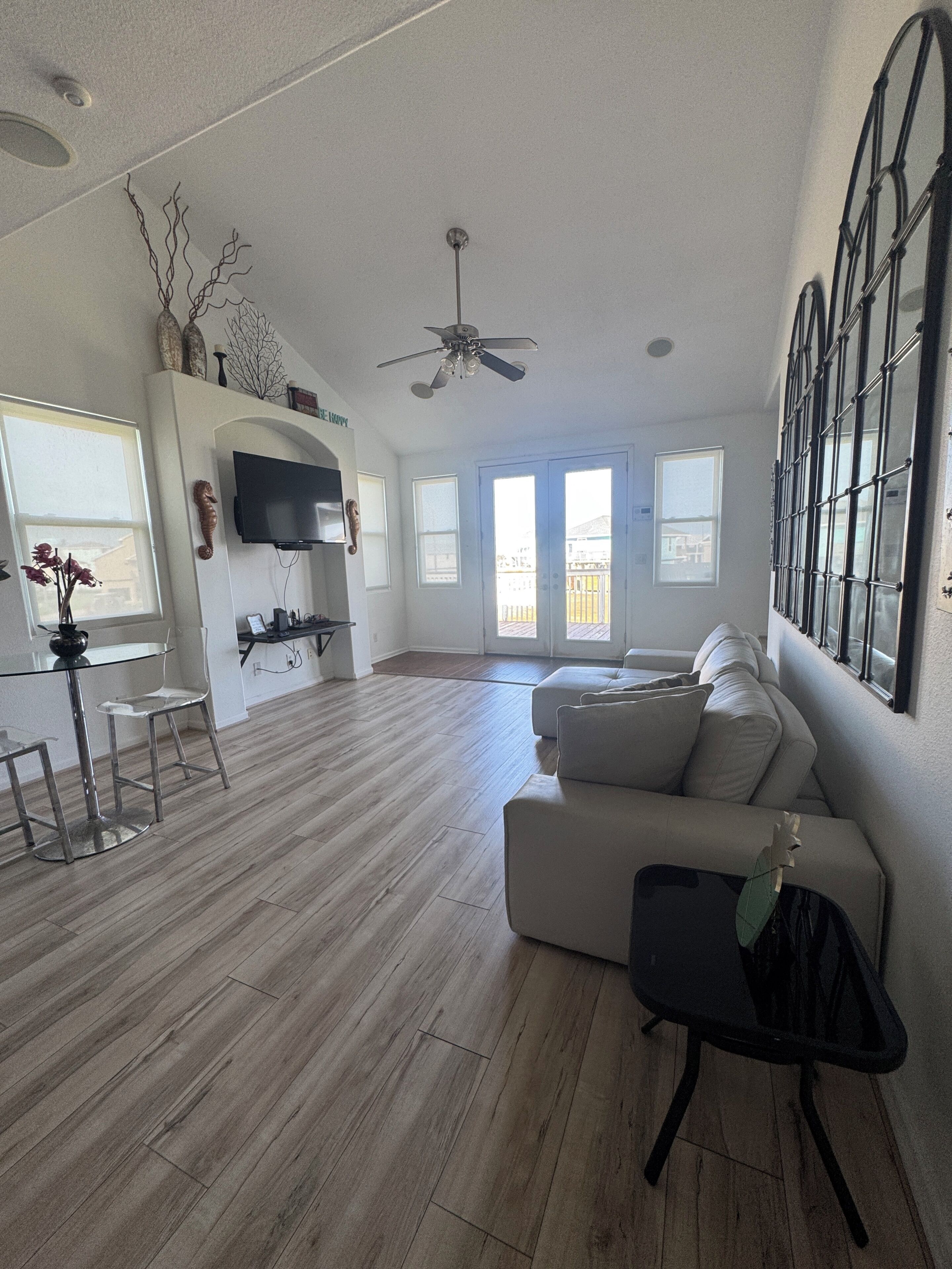A spacious living room with a large window, a ceiling fan, a flat-screen TV, and a seating area with a sofa and a small table.