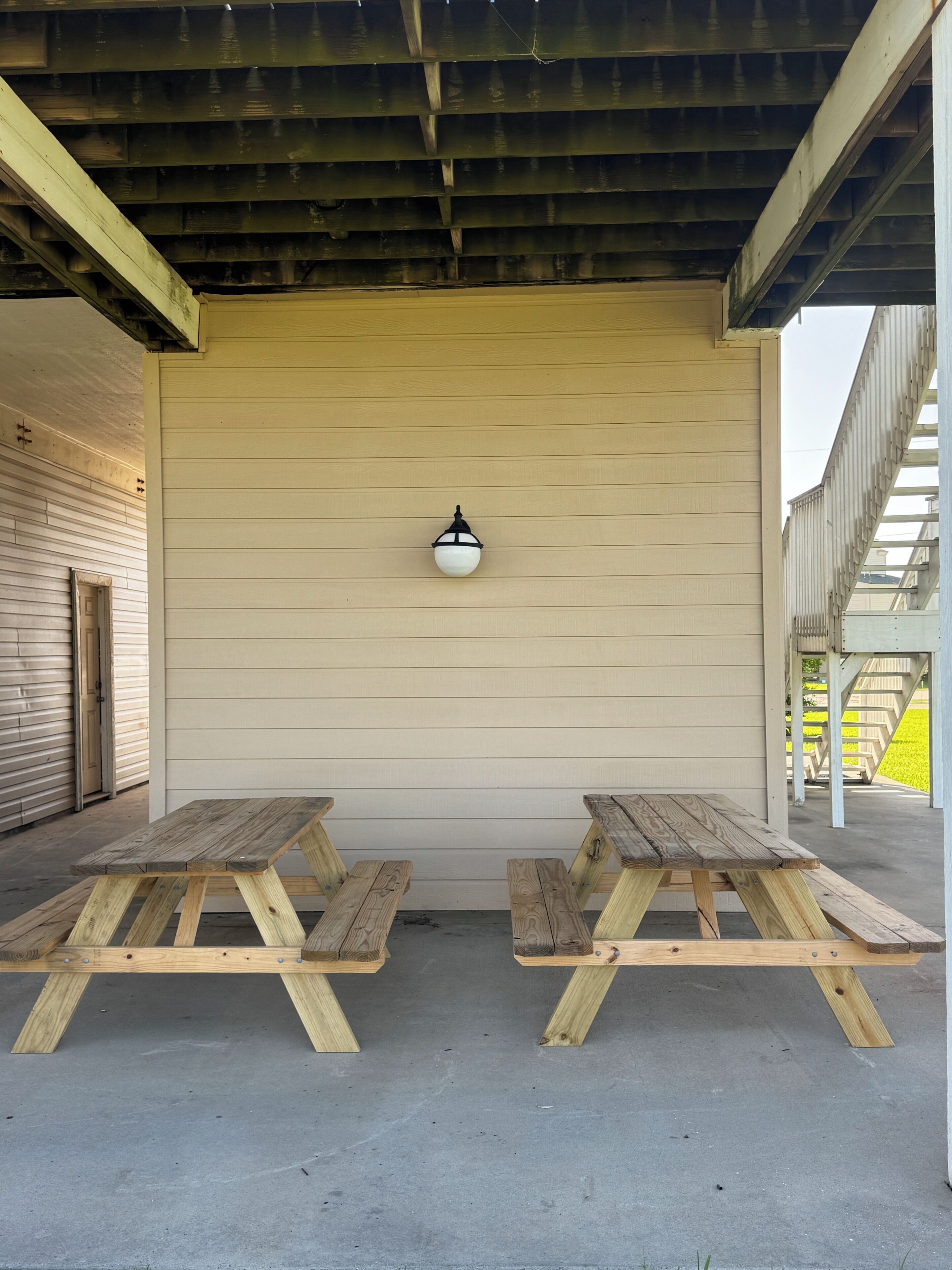 A covered outdoor area with two wooden picnic tables, a light fixture, and a building with horizontal siding.