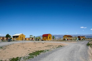 Un camino rural con varios edificios de madera, incluyendo graneros y casas, en un paisaje desértico con montañas al fondo.