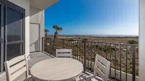 A balcony with a round table and two chairs, overlooking a beach with sparse vegetation and a clear sky.