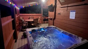 Un bain à remous doté d’une terrasse en bois et illuminé de guirlandes lumineuses, entouré de chaises et d’une table.