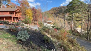 Une cabane rustique dotée d’un porche et d’une petite dépendance située sur une colline, le tout entouré d’un feuillage automnal et surplombant un ciel dégagé.