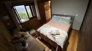 Une chambre avec un plafond en bois, un lit recouvert d’une literie à motifs floraux, une table de chevet ornée d’un vase de fleurs et une fenêtre donnant sur un paysage verdoyant.