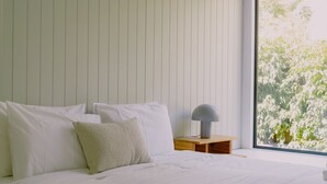 A bedroom with a bed, pillows, a bedside table with a lamp, and a window with a view of greenery. - Monument House (Daylesford)
