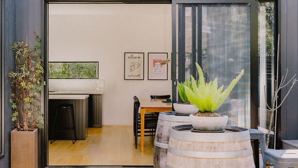 A modern kitchen with a large window, a dining area with a wooden table and chairs, and potted plants. - Monument House (Daylesford)