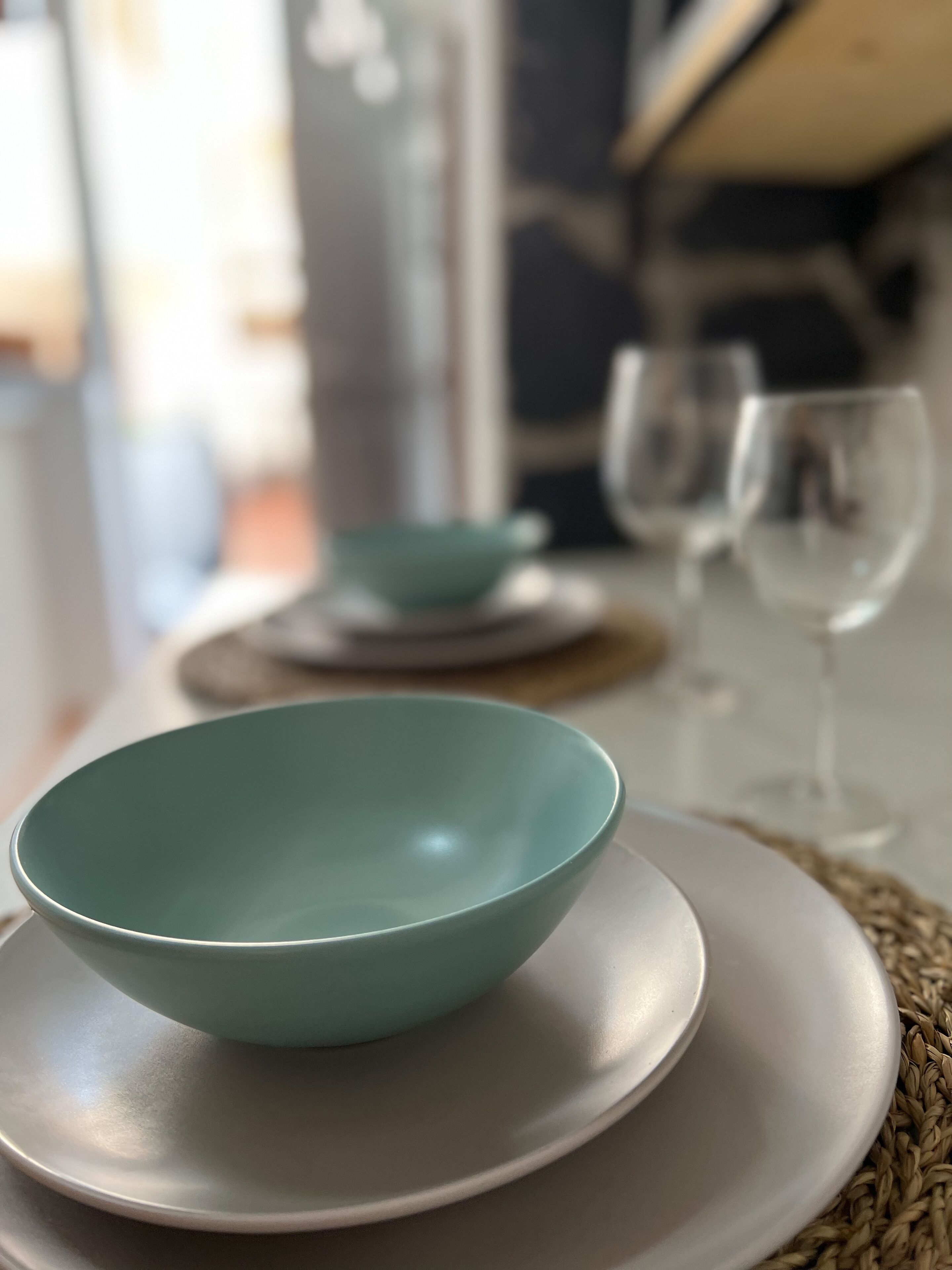 A dining table with a teal bowl on a white plate, a smaller bowl on a coaster, and two wine glasses in the background.