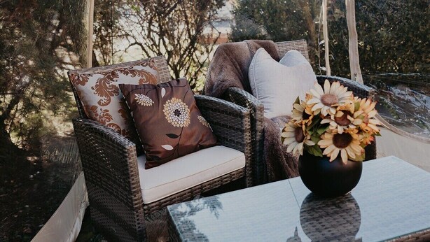Une chaise en osier avec un coussin et des oreillers à motifs, une table en verre ornée d’un vase de tournesols, et une vue sur des arbres et un bâtiment en arrière-plan.