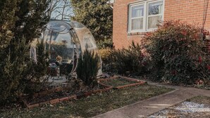Une serre dotée d’un toit transparent, entourée d’un jardin où poussent différentes espèces de plantes, avec une maison en briques en arrière-plan.