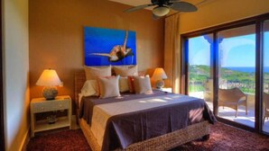 A bedroom with a bed, two bedside tables with lamps, a ceiling fan, a large window with a view, and a painting on the wall. - Beautiful Golf Villa 2 Puerto Los Cabos (Puerto Los Cabos)