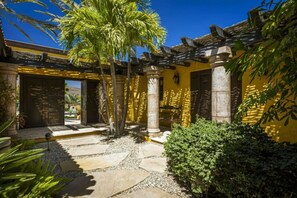 A courtyard with a stone pathway, palm trees, and a yellow building with columns.