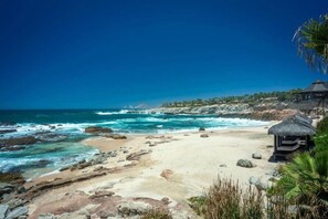 A beach with a thatched-roof structure, rocky shoreline, and clear blue sea.