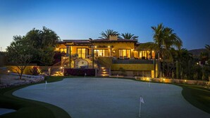 Une grande maison bien éclairée, avec un terrain de golf devant, et un escalier menant à un balcon.