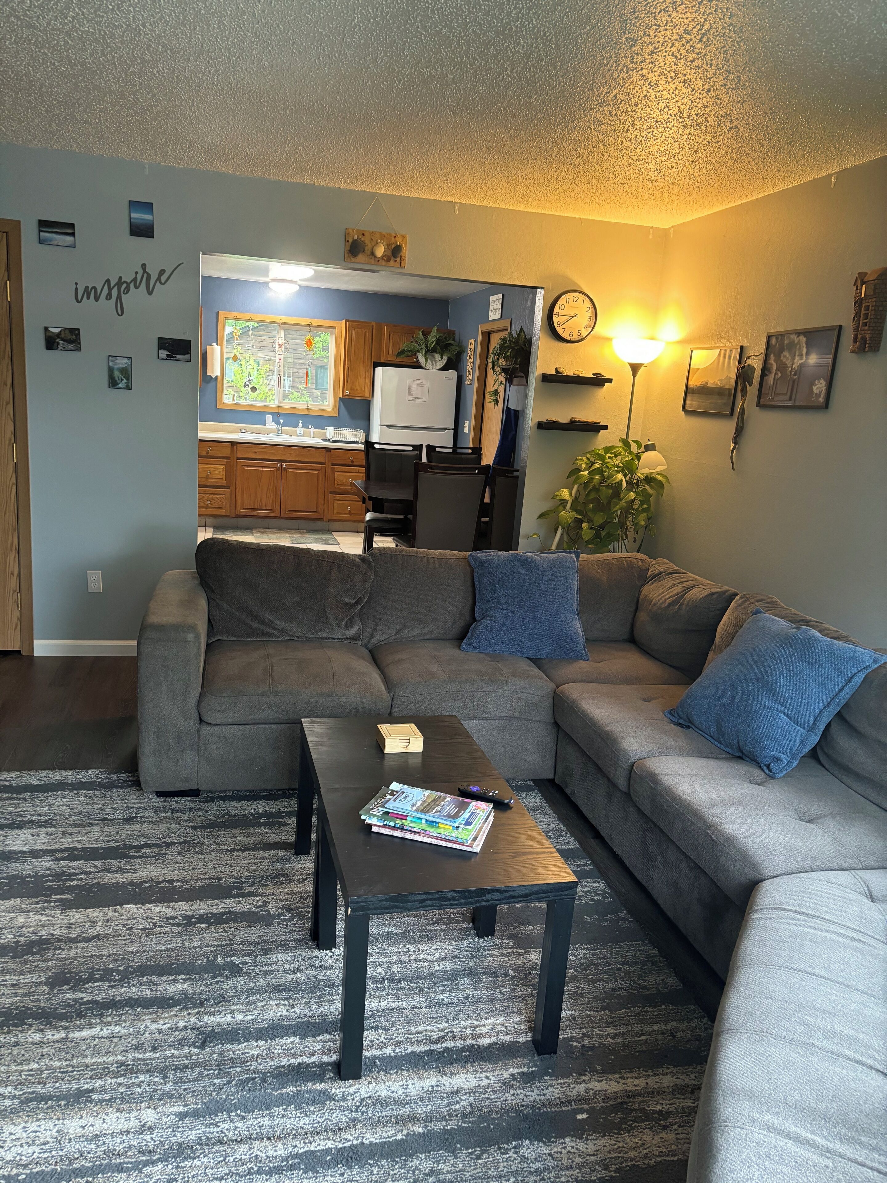 A living room with a sectional sofa, a coffee table with books and a remote, a kitchen in the background, and a wall with a clock and pictures.