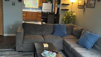 A living room with a sectional sofa, a coffee table with books and a remote, a kitchen in the background, and a wall with a clock and pictures.