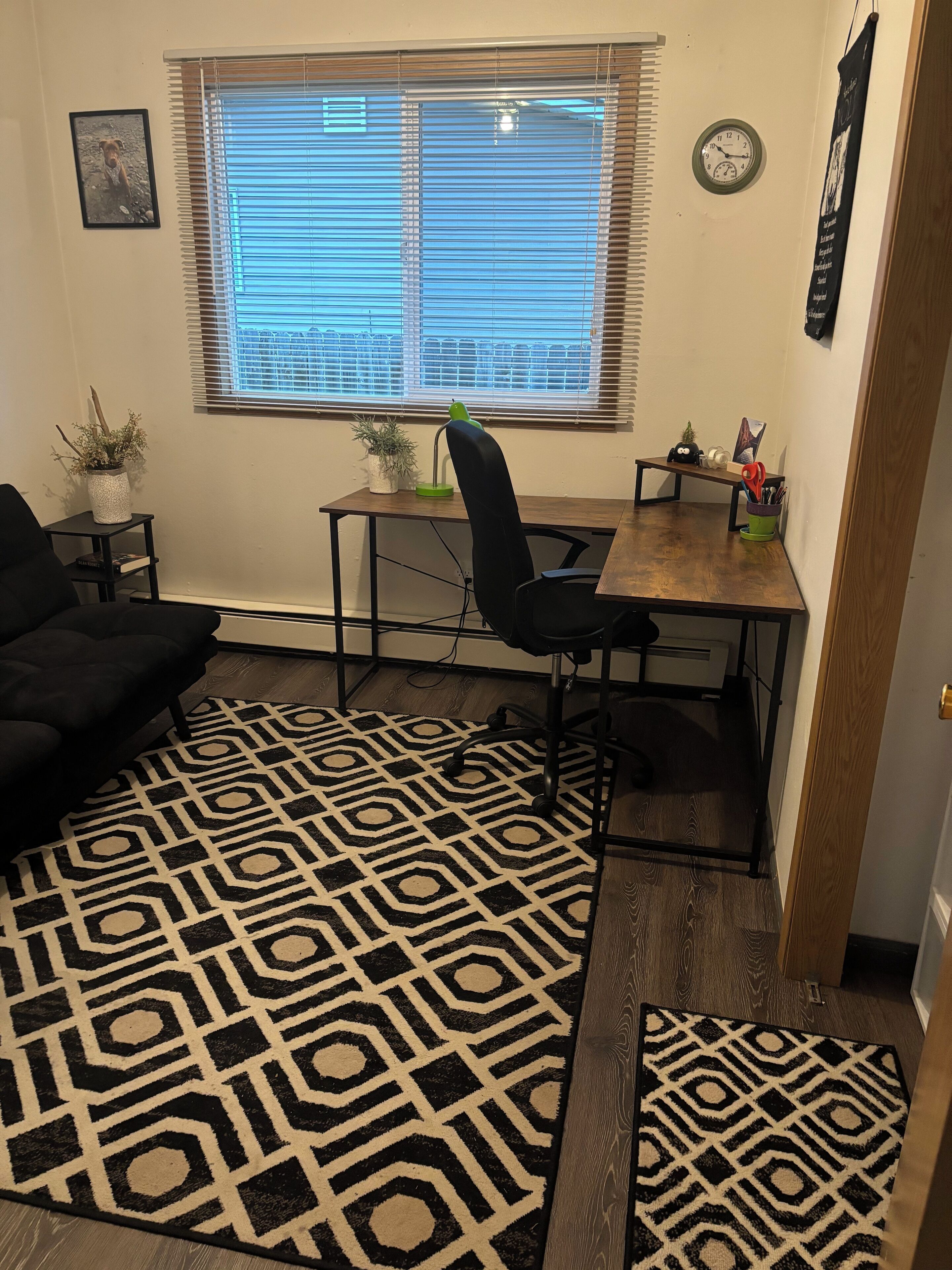 Una habitación con un escritorio, dos sillas, un tapete estampado, una planta pequeña en maceta y un reloj en la pared.