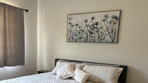 A bedroom with a large bed, a nightstand, a wall-mounted painting, and a window with curtains.