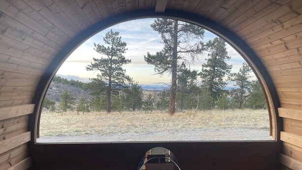 A circular wooden cabin with a large arched window offering a view of a forest and mountains. Inside, there are wooden benches and a metal chair. - Amazing  Views--Spacious 3BR, outdoor barrel sauna, dog friendly (Hartsel)