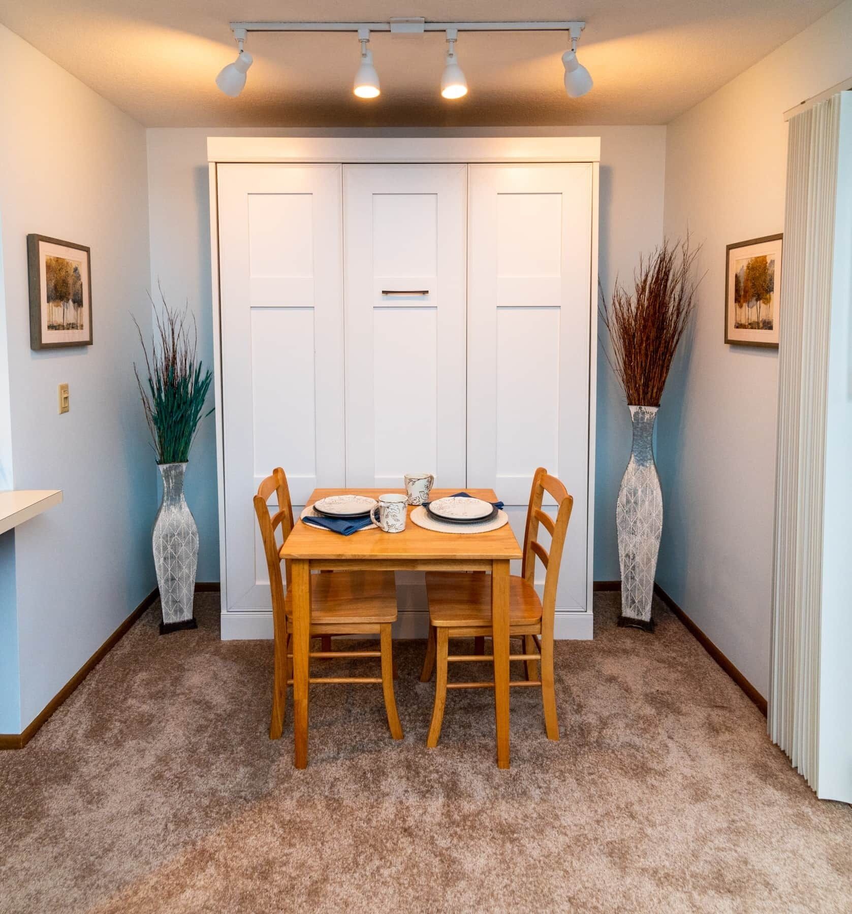 Un coin repas avec une table dressée pour quatre personnes, flanqué de deux portes blanches et de deux vases ornés de plantes.