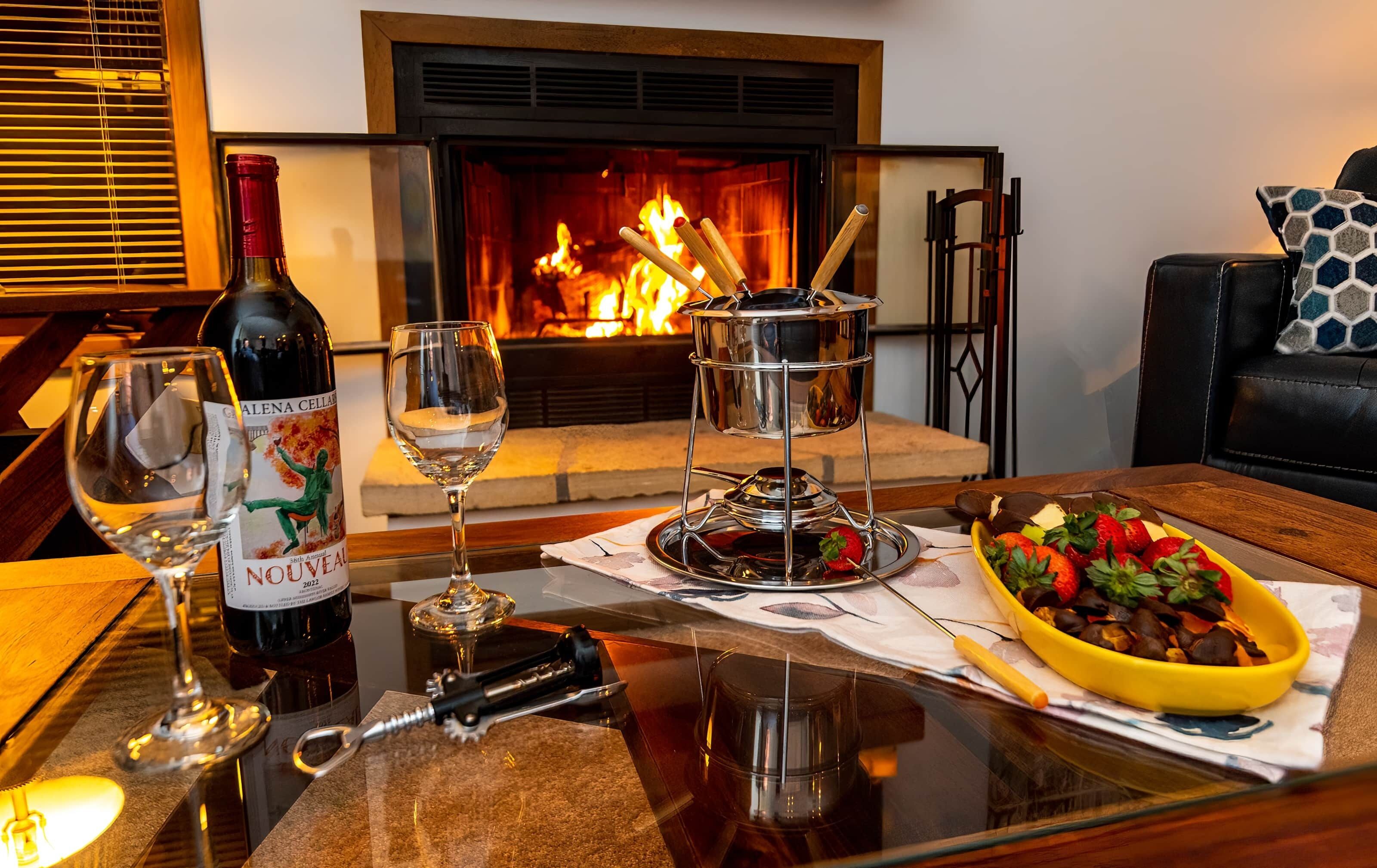 A table with a bottle of wine, a glass, a candle, a fruit bowl, and a candle holder in front of a fireplace.