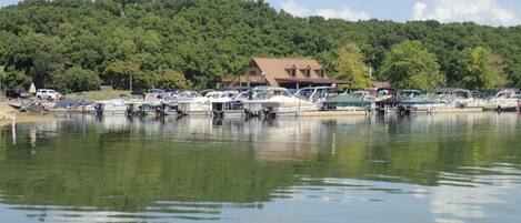 A marina with boats docked, a building with a sloped roof, and a forested hill in the background.