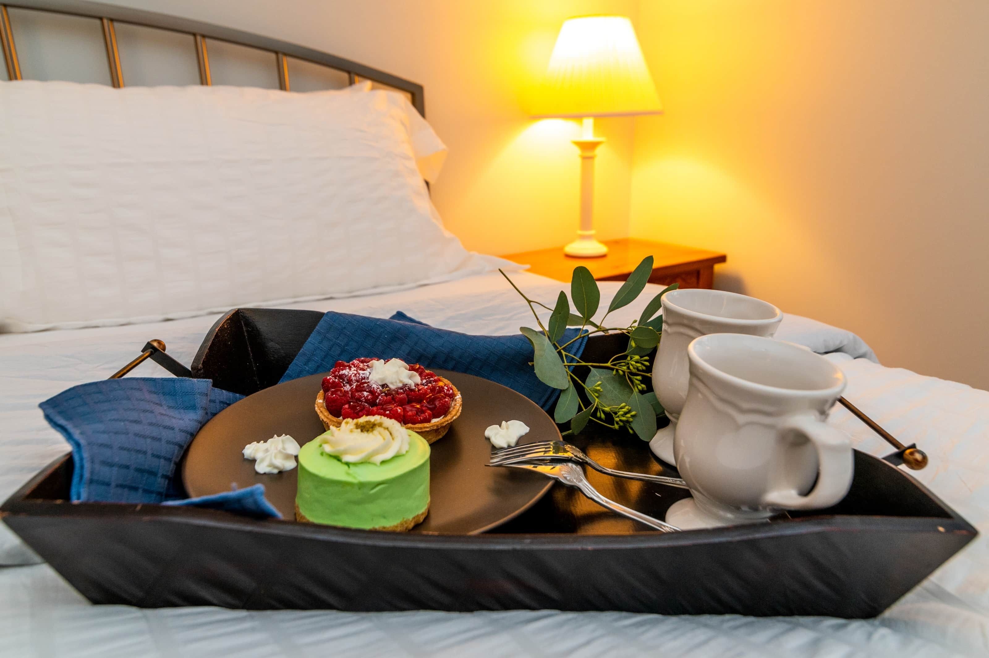 Ein Tablett mit Desserts und einer Tasse auf einem Bett, im Hintergrund ein Nachttisch und eine Lampe.
