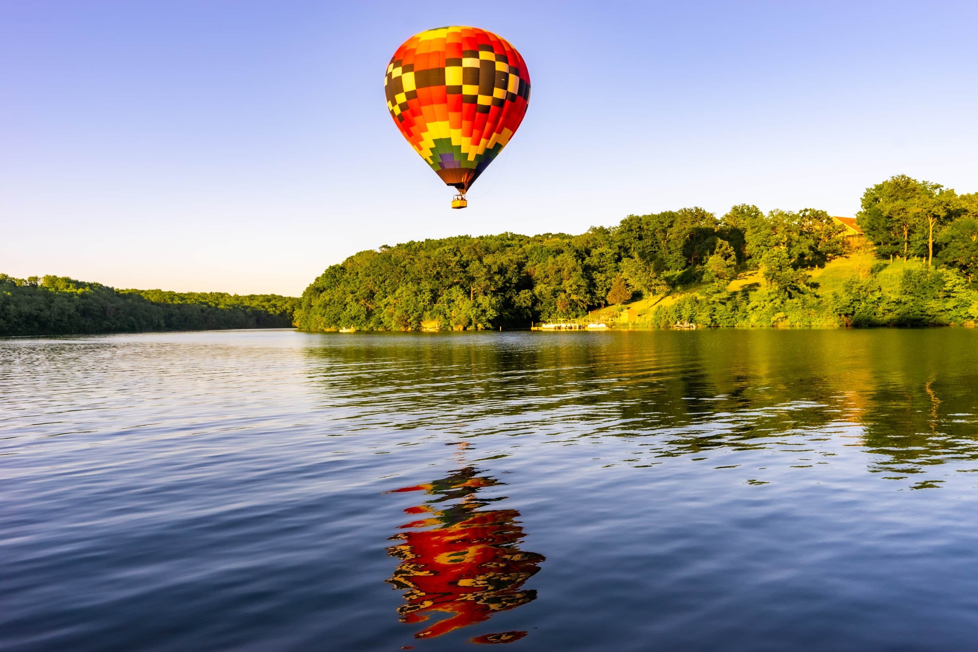 A hot air balloon over a calm lake with a forested hillside in the background.