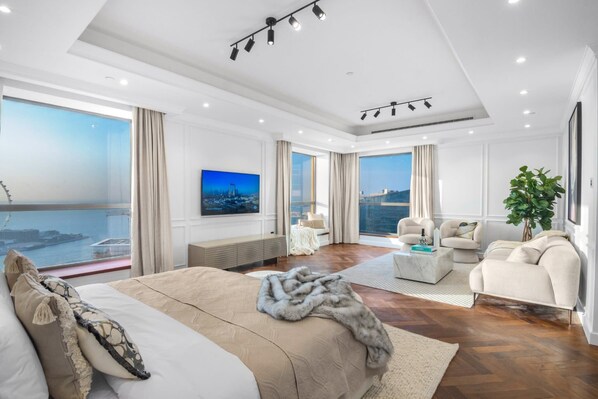 A modern hotel room with a large bed, a seating area with a sofa and armchairs, a large window with a city view, and a flat-screen TV mounted on the wall.