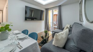 Una sala de estar moderna con un sofá gris, una mesa de comedor preparada para cuatro comensales y un televisor grande montado en la pared.